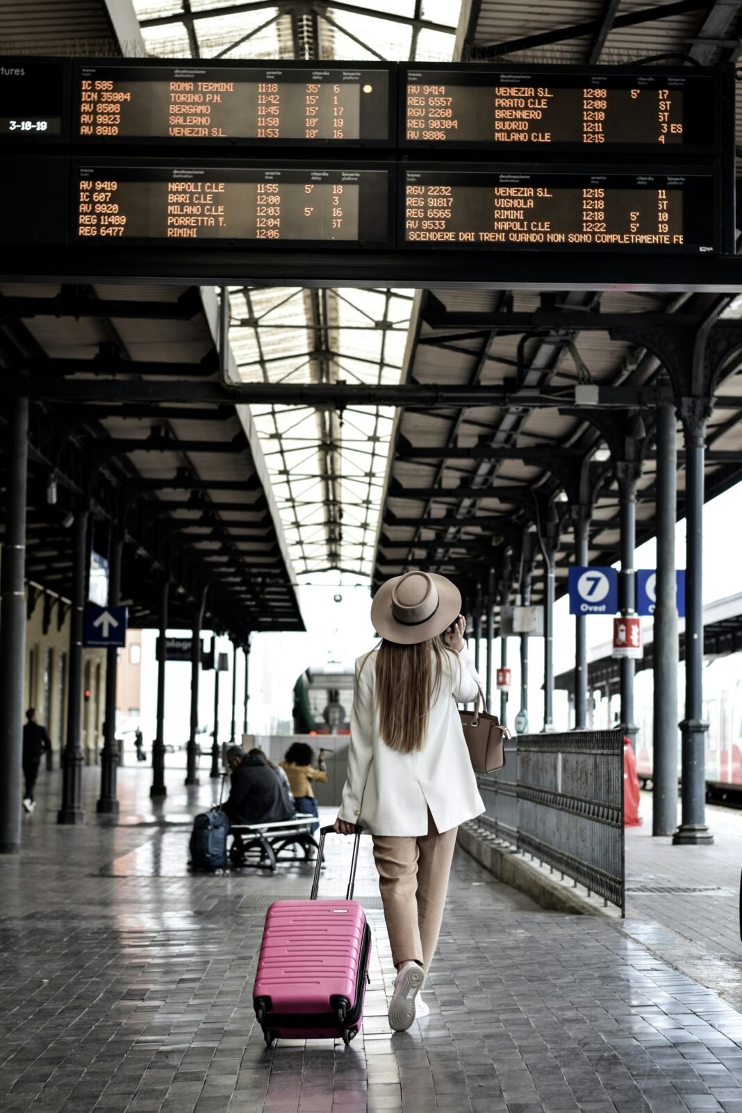Mulher caminhando por um corredor de aeroporto, segurando uma mala de viagem cor de rosa, com luz natural entrando pelas janelas.
