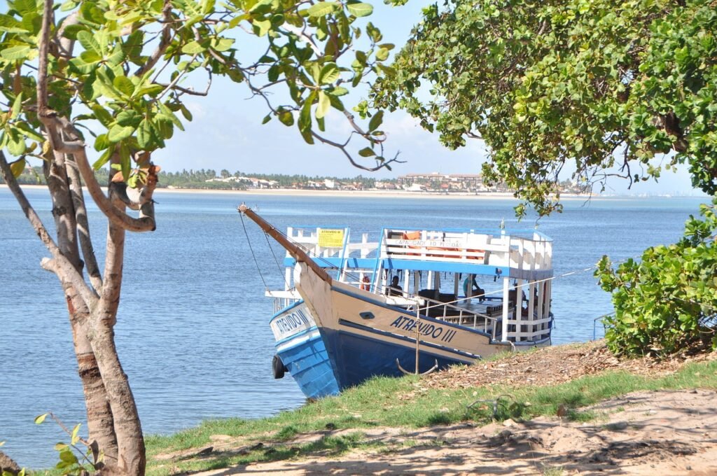 Barco de madeira encostado na areia com o mar azul ao fundo, em uma praia de Maceió, Alagoas.