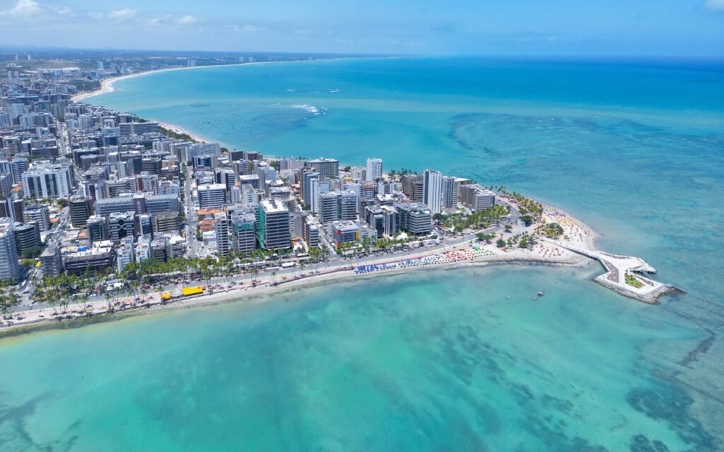 Vista panorâmica da cidade de Maceió com edifícios à beira-mar e o oceano azul ao fundo sob céu ensolarado.