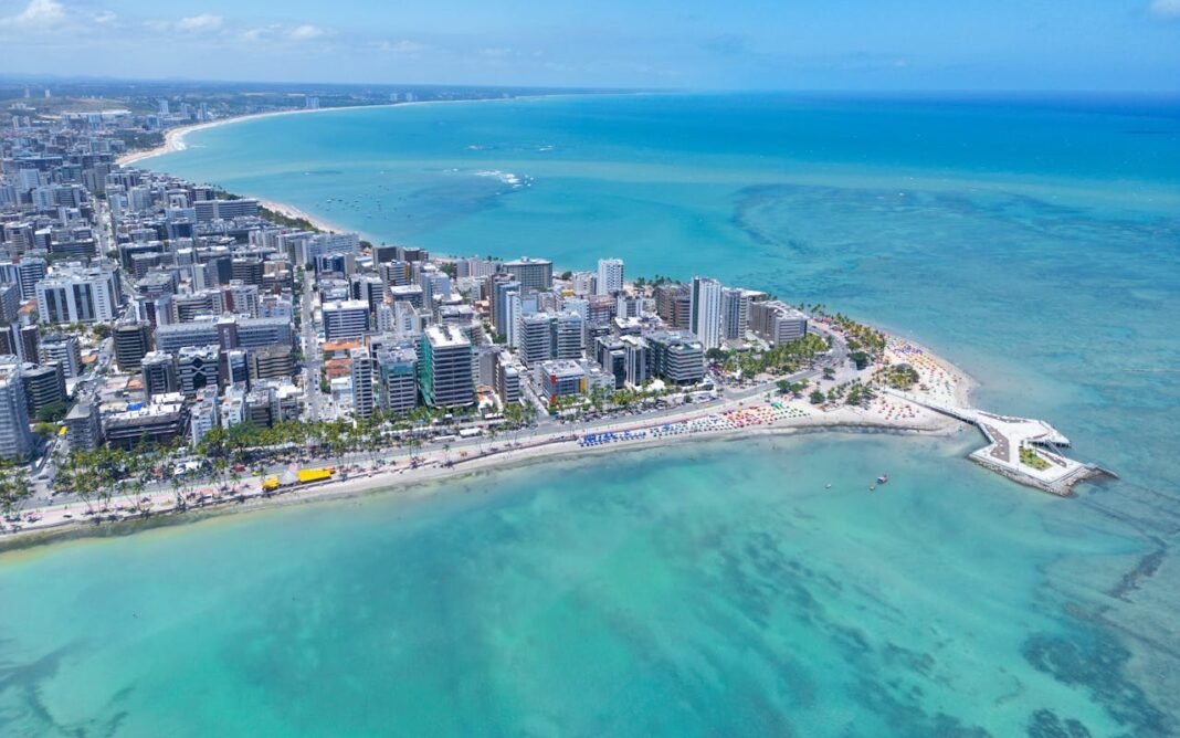 Vista panorâmica da cidade de Maceió com edifícios à beira-mar e o oceano azul ao fundo sob céu ensolarado.