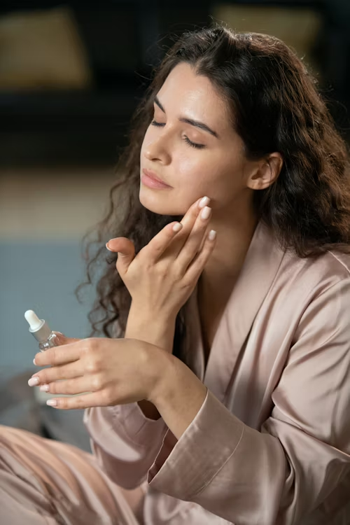 Mulher sentada com as pernas cruzadas, usando pijama de cetim e aplicando sérum facial com as mãos.