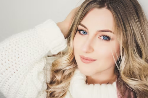 Mulher loira de olhos azuis usando maquiagem neutra e blusa branca, com expressão serena e aparência natural.