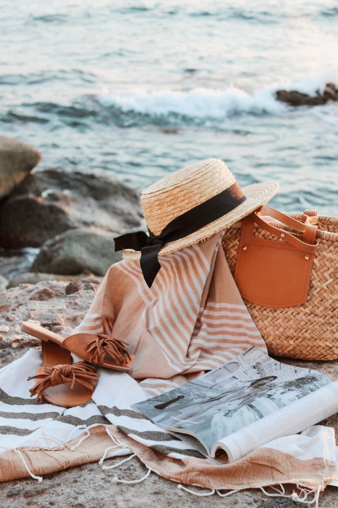 Bolsas tressê, chapéu de palha, sandália e revista dispostos sobre a areia da praia em uma composição de verão.