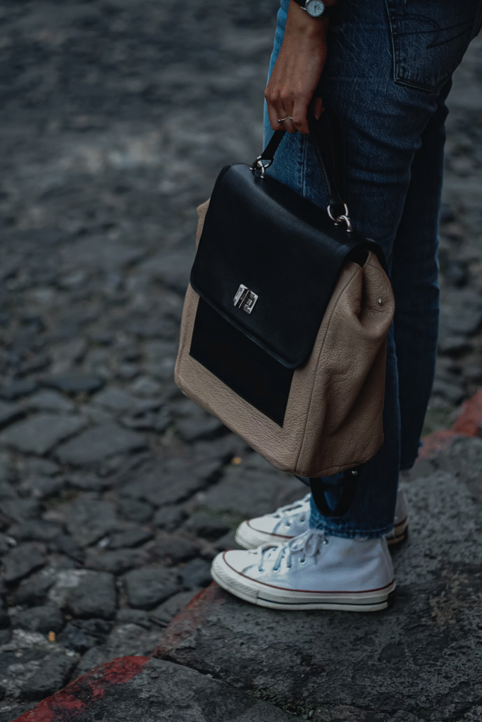 Mulher parada em uma calçada usando calça jeans e tênis branco, segurando uma bolsa marrom e preta.