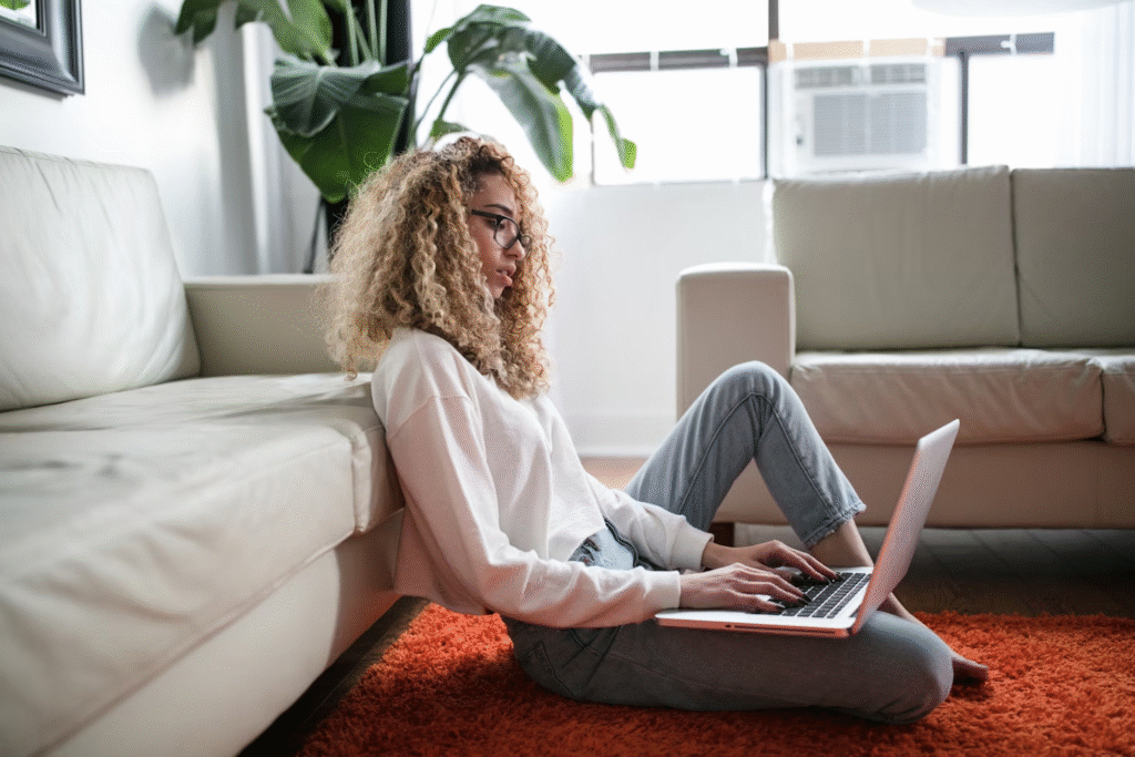 Mulher trabalha em seu notebook sentada em um tapete vermelho, encostada em um sofá branco.