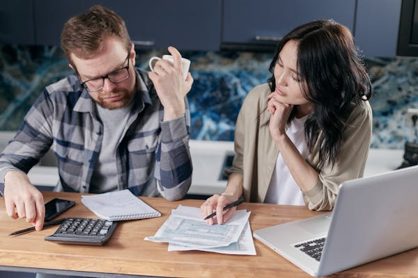 Casal sentado à mesa com computador, calculadora e papéis, concentrados em organização das suas finanças