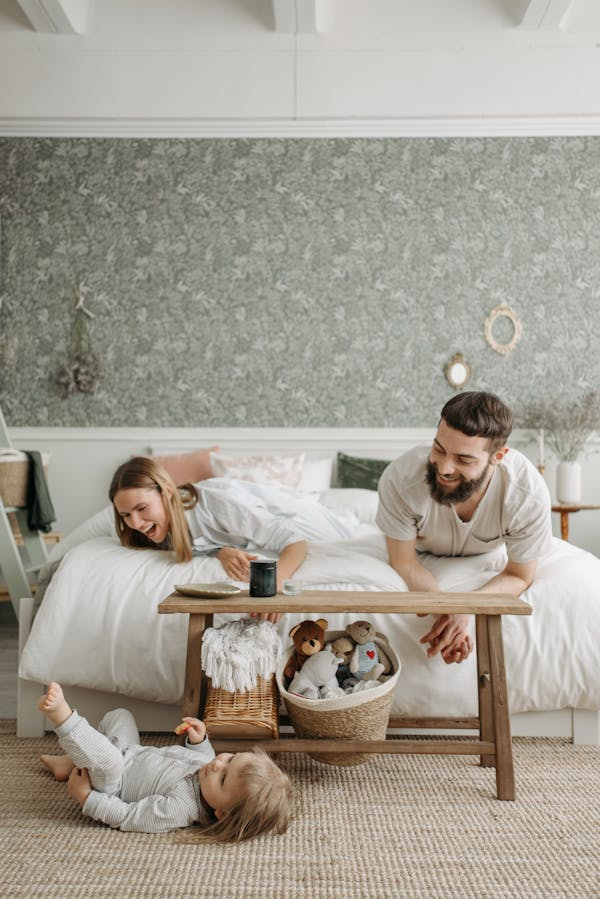 Casal deitado de bruços na cama observa e interage com o filho que brinca no chão do quarto.