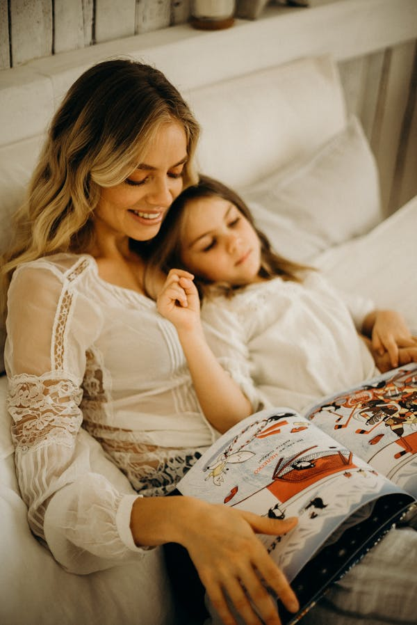 Mãe e filha deitadas na cama, abraçadas, lendo juntas um livro de historinhas.