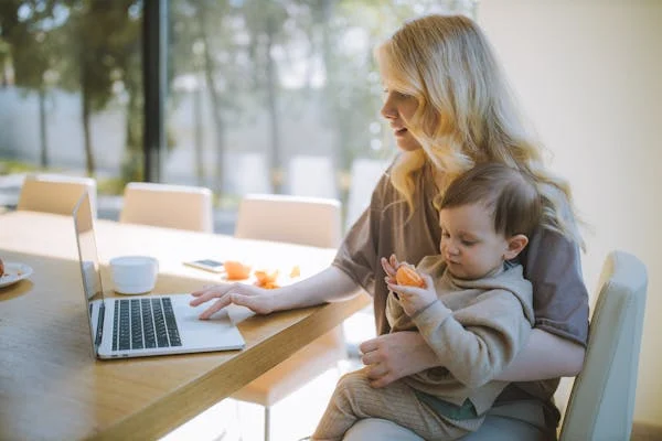 Mulher sentada trabalha no notebook enquanto segura o filho pequeno que come uma laranja.
