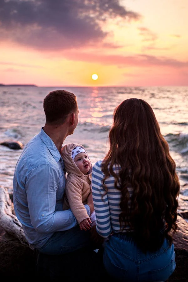 Casal sentado de frente para o mar com o filho no colo, observando juntos o pôr do sol.