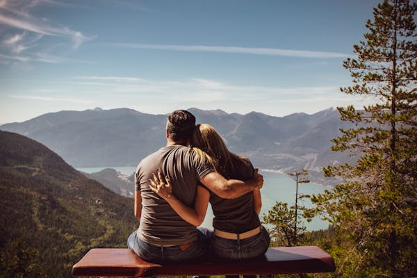 Casal sentado abraçado no topo de uma montanha, contemplando o mar e a paisagem montanhosa ao redor.