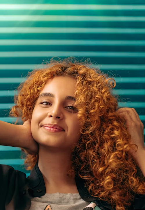 Mulher ruiva de cabelos cacheados posa com as mãos no cabelo em frente a uma parede azul.