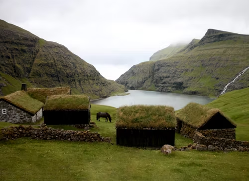 Paisagem das Ilhas Faroé com casas tradicionais de telhados cobertos de grama verde, cercadas por montanhas e vegetação típica da região.