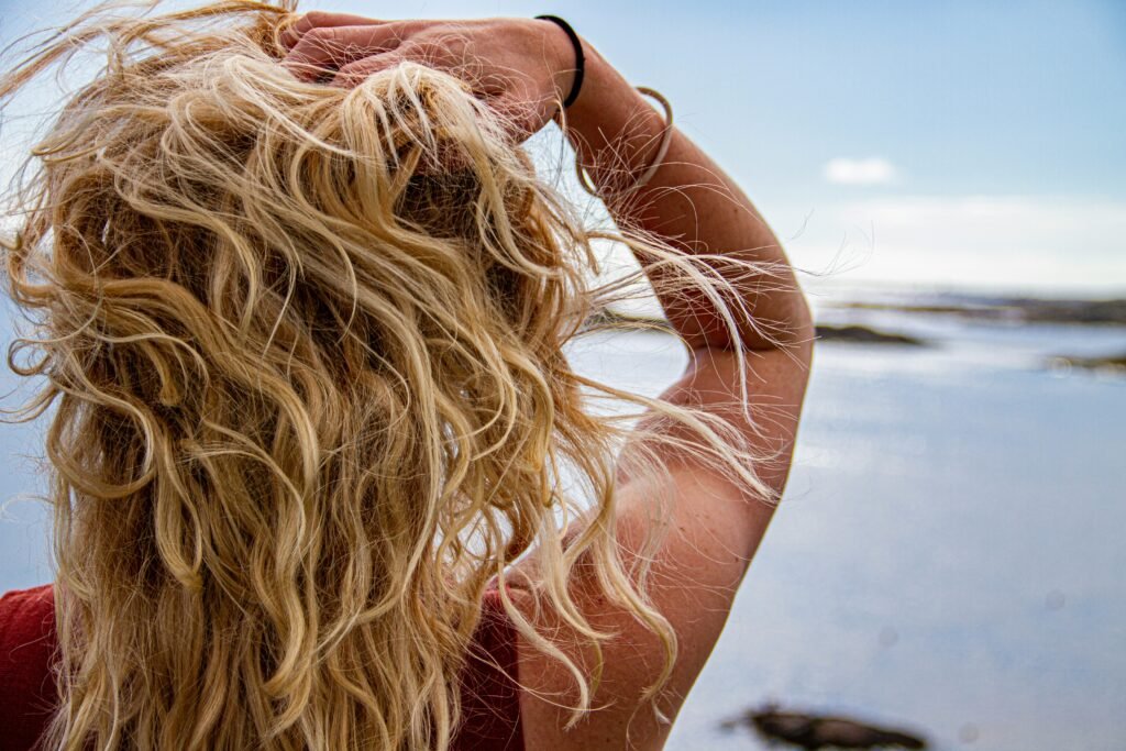 Mulher loira vista de costas, com uma das mãos no cabelo, olhando para o mar em um cenário tranquilo e ensolarado.