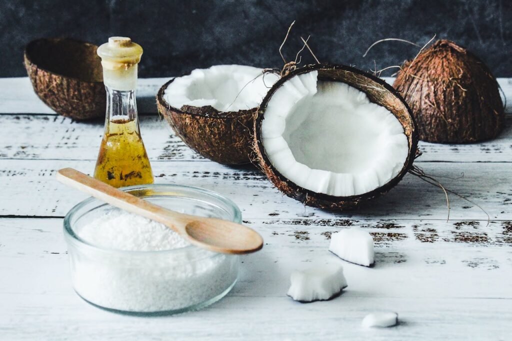 Cocos secos partidos ao meio ao lado de uma jarra com óleo de coco sobre uma mesa rústica de madeira.