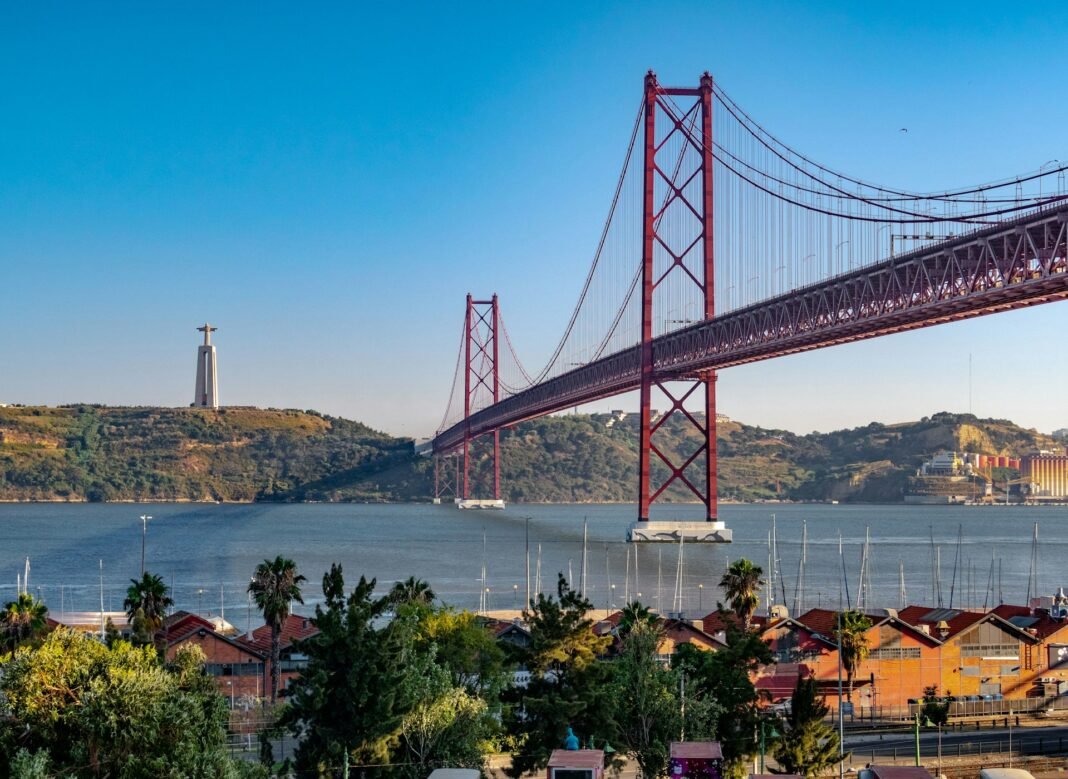 Ponte 25 de Abril em Lisboa em um dia ensolarado, com céu azul e o rio Tejo refletindo a luz do sol.