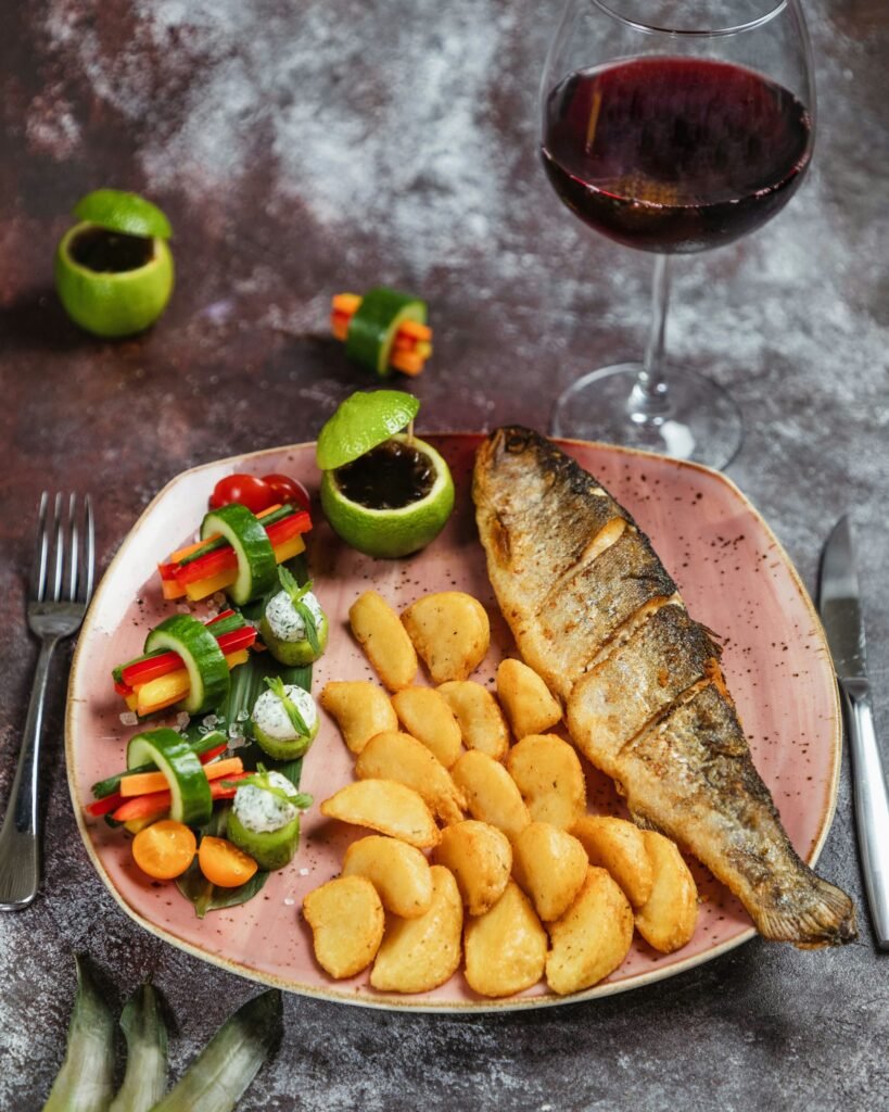 Prato com sardinha frita acompanhada de batatas, salada colorida e uma taça de vinho ao lado.