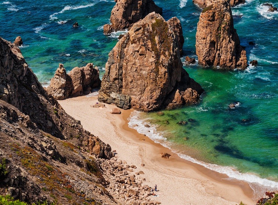 Praia de Sintra em Lisboa com águas azuis cristalinas, rochas e paisagem natural preservada em um dia claro.