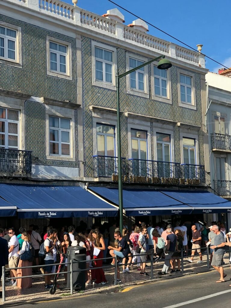 Fachada da Pastelaria Pastéis de Belém em Lisboa, com toldos azuis e pessoas aguardando na fila para comprar pastéis de belém.